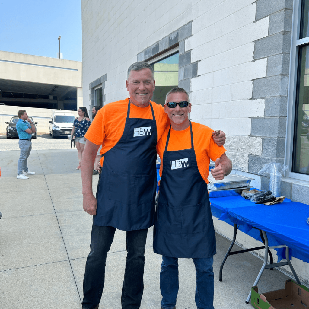 Two men, John Leach and Phil Scott, smiling at a cookout, wearing blue HBW Construction aprons. 